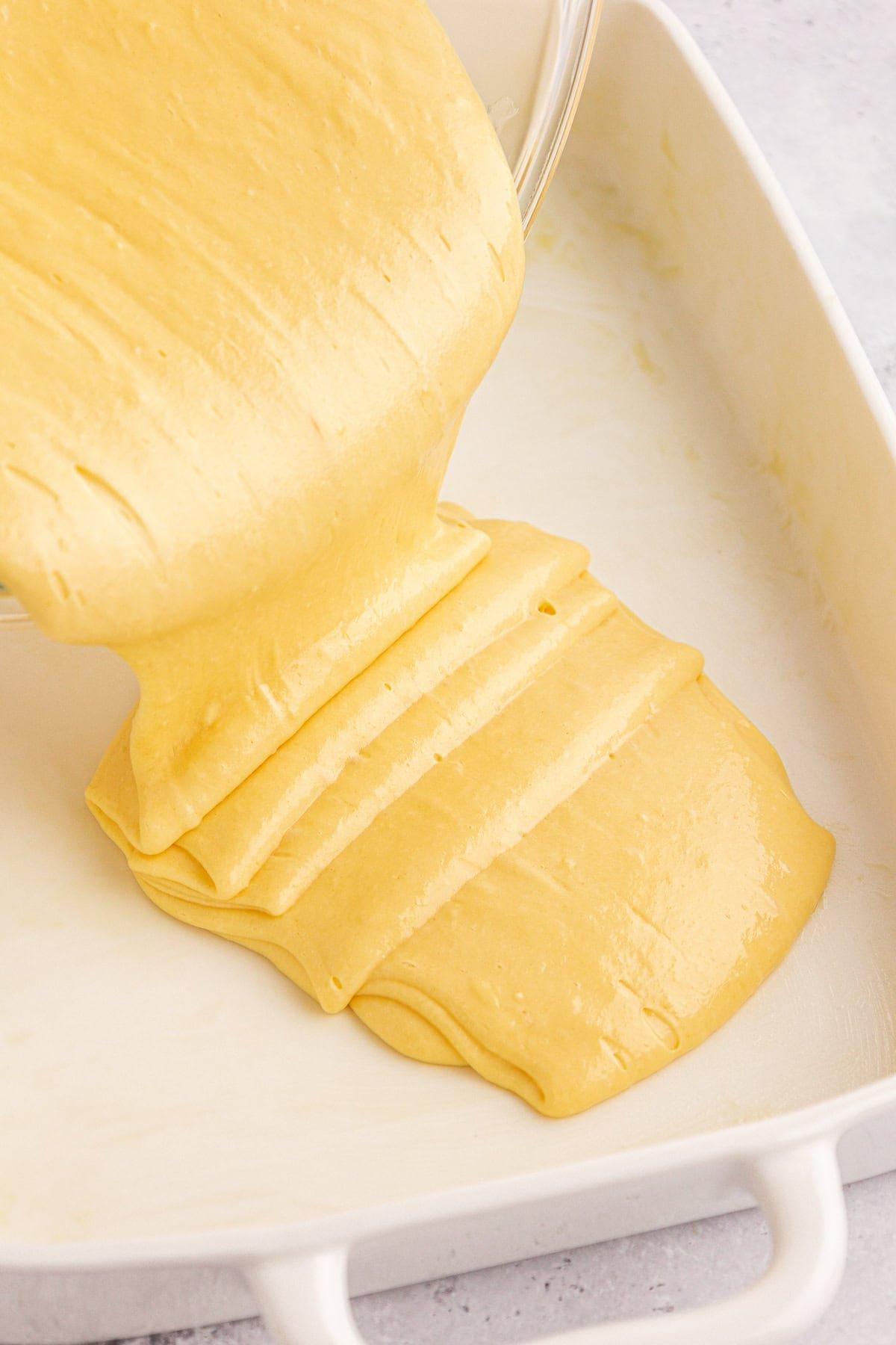 L'impasto giallo della torta viene versato da una ciotola in una teglia rettangolare bianca, destinata a trasformarsi in una deliziosa torta Honey Bun. L'impasto liscio e lucido si stende uniformemente sul fondo del piatto, promettendo una dolce perfezione.