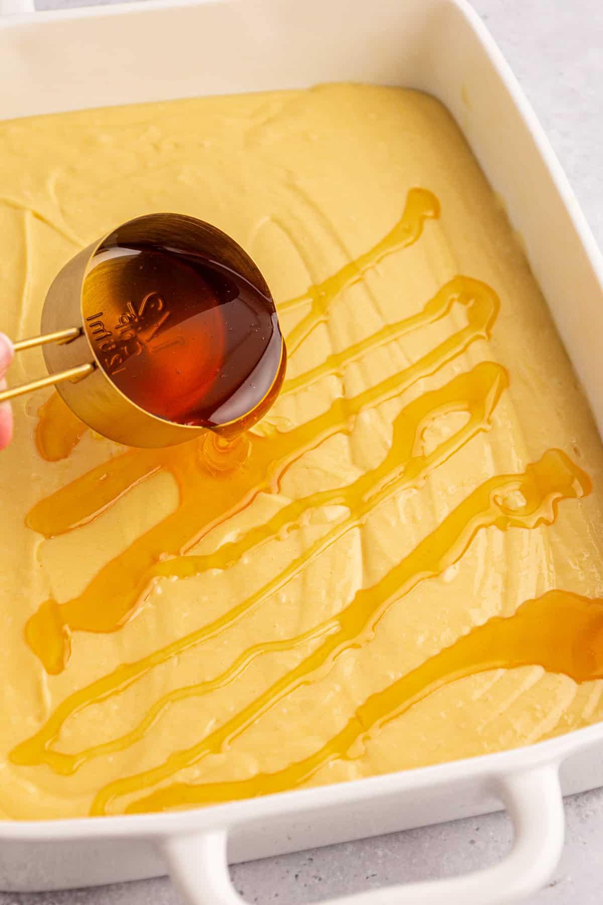 Una teglia da forno riempita con impasto marrone chiaro attende la sua trasformazione in una deliziosa torta Honey Bun. Una mano versa miele da una tazza da 1/2 tazza a zig-zag sopra la miscela. La teglia, pronta per il forno, è invitante sul piano di lavoro di colore chiaro.