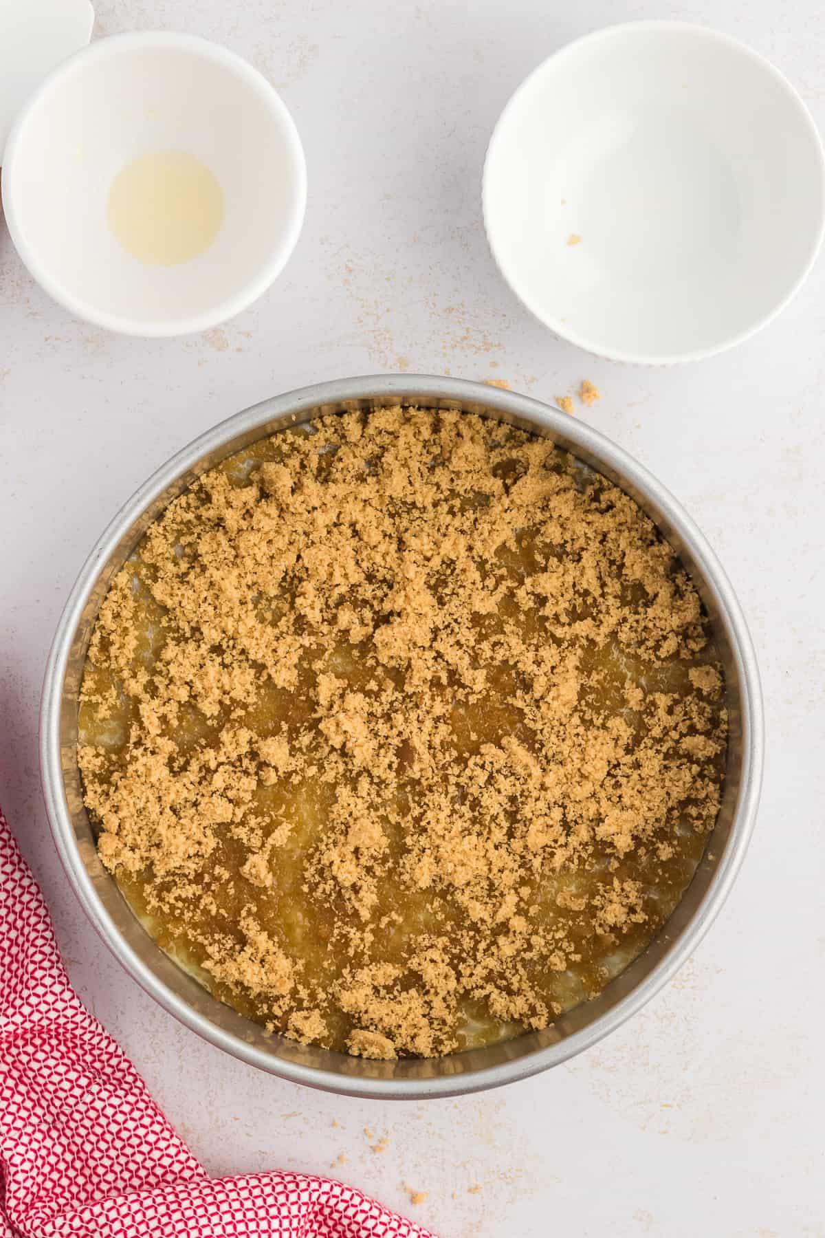 Una teglia rotonda per torte con burro fuso e zucchero di canna nel fondo, circondata da due piccole ciotole bianche vuote e un canovaccio rosso e bianco.