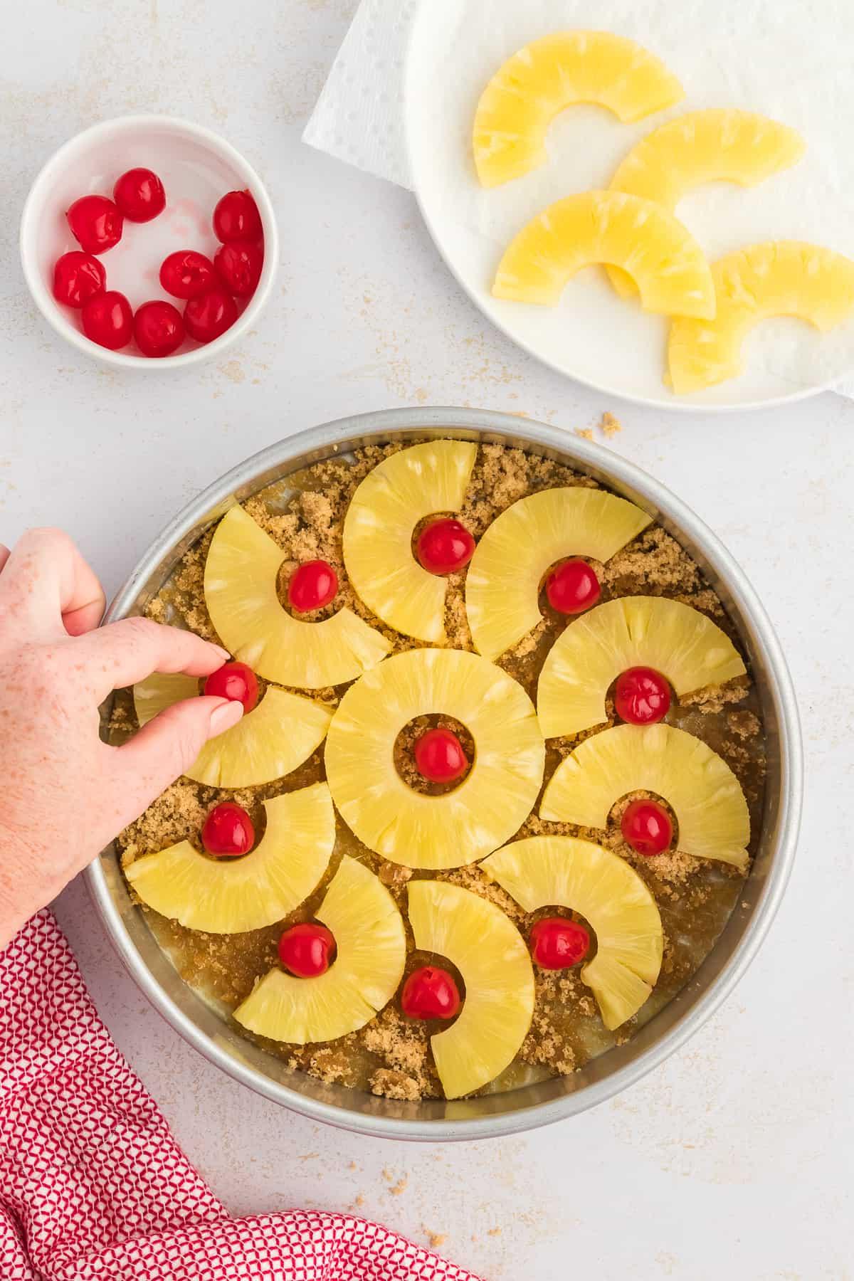 Una teglia per torte con zucchero di canna e burro fuso nel fondo e una mano che sistema fette di ananas e ciliegie al maraschino, accanto a un canovaccio rosso e bianco, un piatto di ananas a fette e una piccola ciotola di ciliegie.