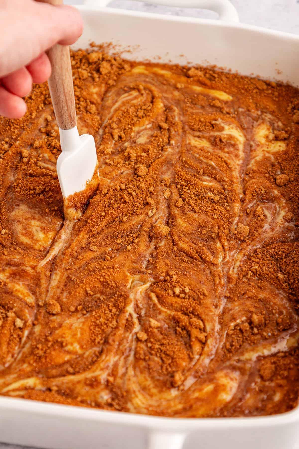 Una mano muove delicatamente una spatola bianca nell'impasto della torta Honey Bun, composto da cannella, zucchero di canna e impasto, in una teglia da forno bianca, creando un bellissimo effetto marmorizzato.
