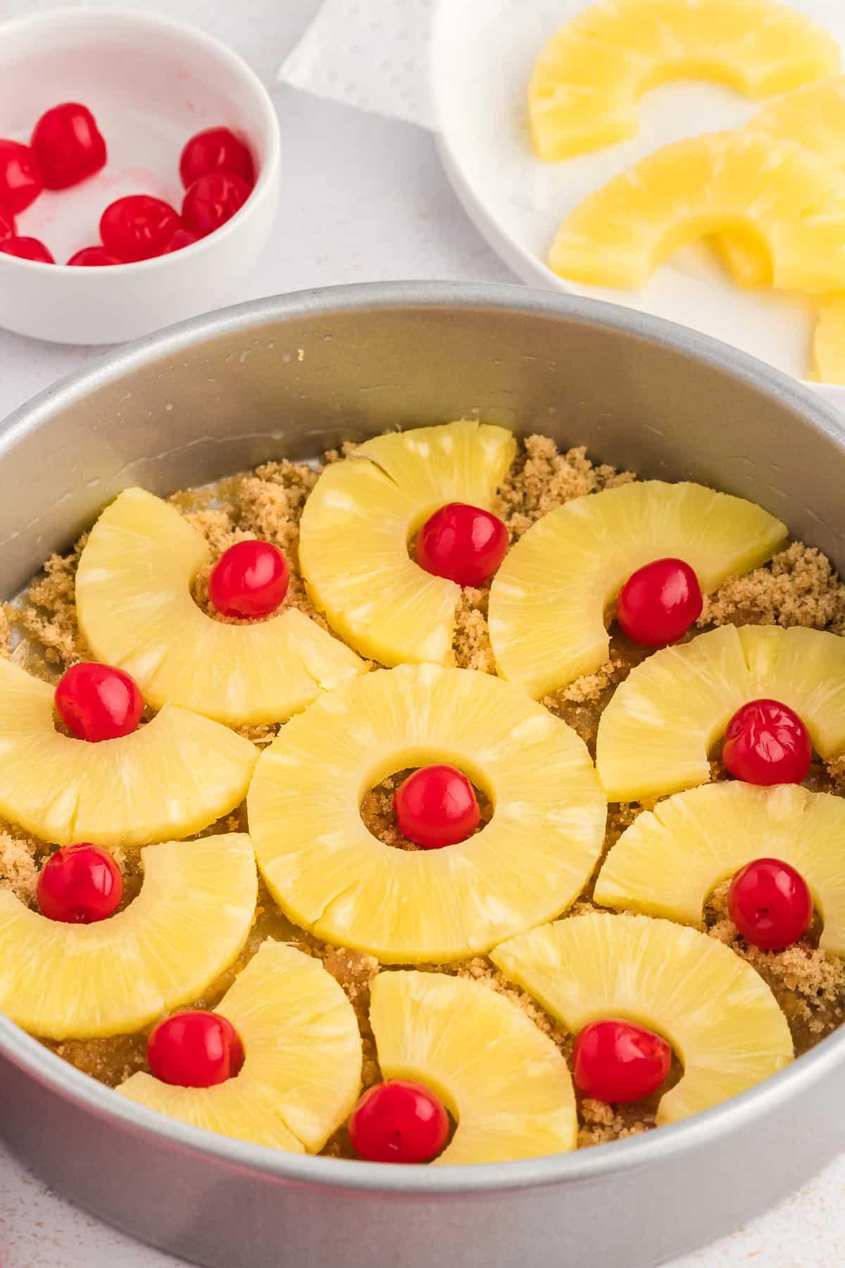 Una teglia per torte con zucchero di canna e burro fuso nel fondo e fette di ananas e ciliegie al maraschino disposte, accanto a un canovaccio rosso e bianco, un piatto di ananas a fette e una piccola ciotola di ciliegie.