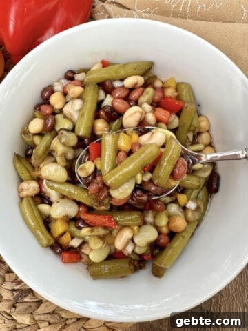 A bowl of green bean salad with a serving spoon, a napkin on the side and peppers near the bowl.
