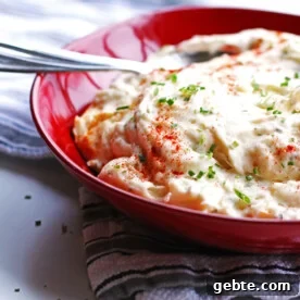 Baked mashed potatoes in a red bowl with a spoon. The potatoes are garnished with chives.