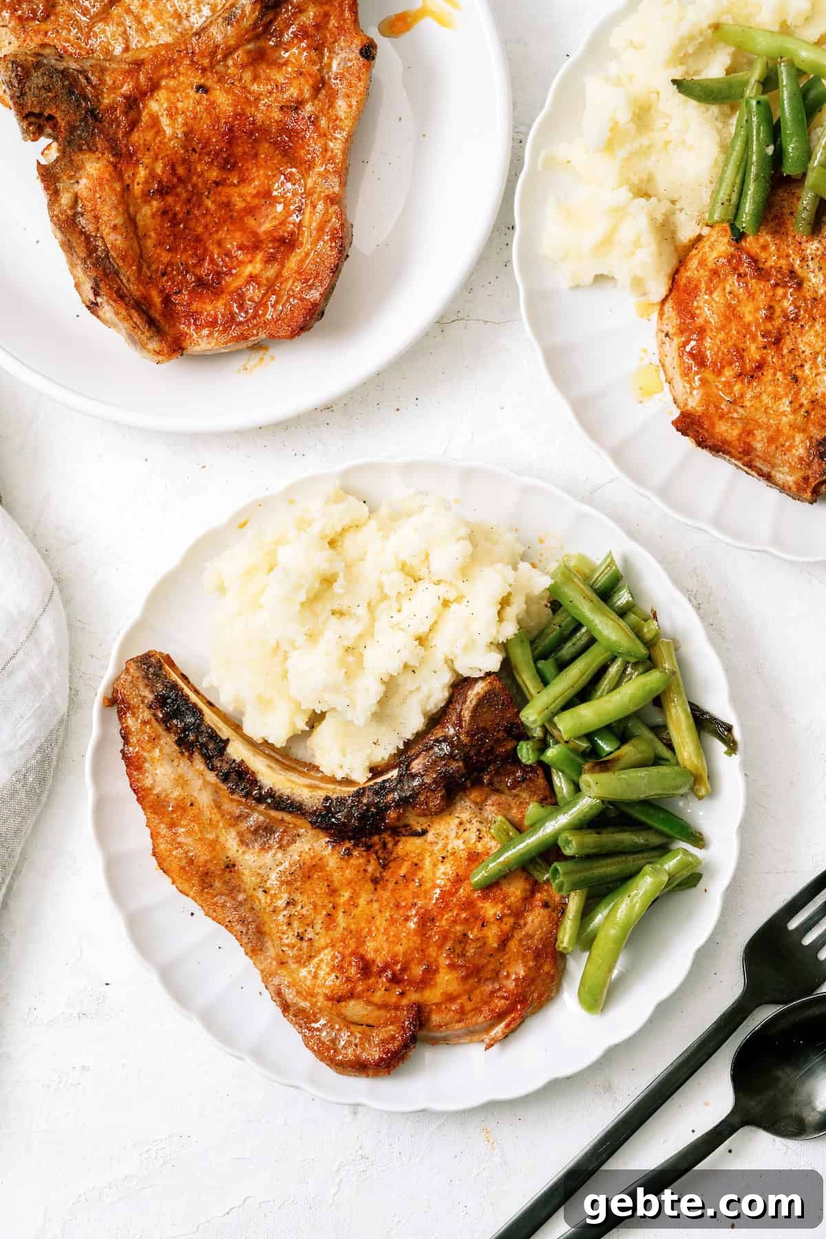 A white plate with a cooked pork chop, mashed potatoes, and green beans. Two additional plates with similar servings are partially visible.