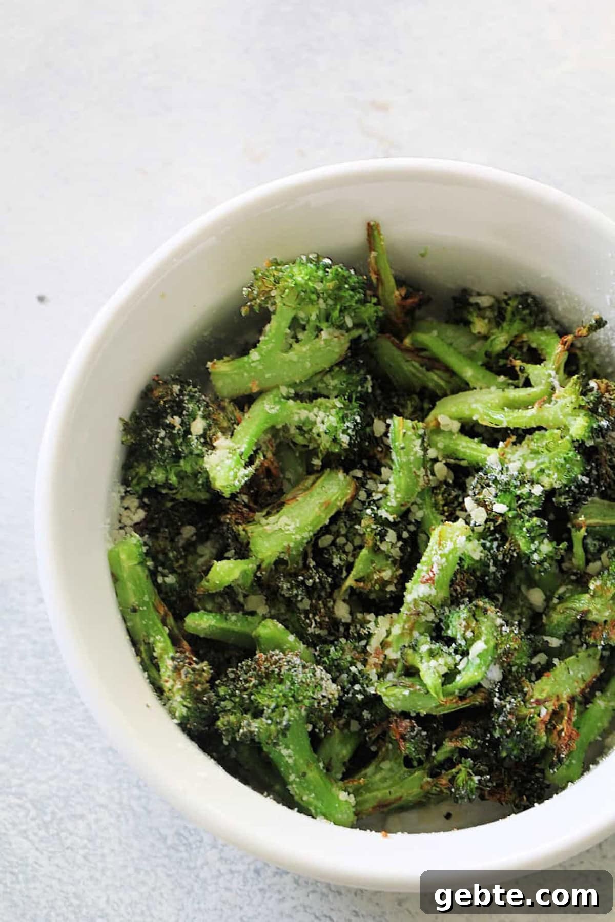 A white bowl filled with roasted broccoli florets topped with grated cheese, set on a light-colored surface.