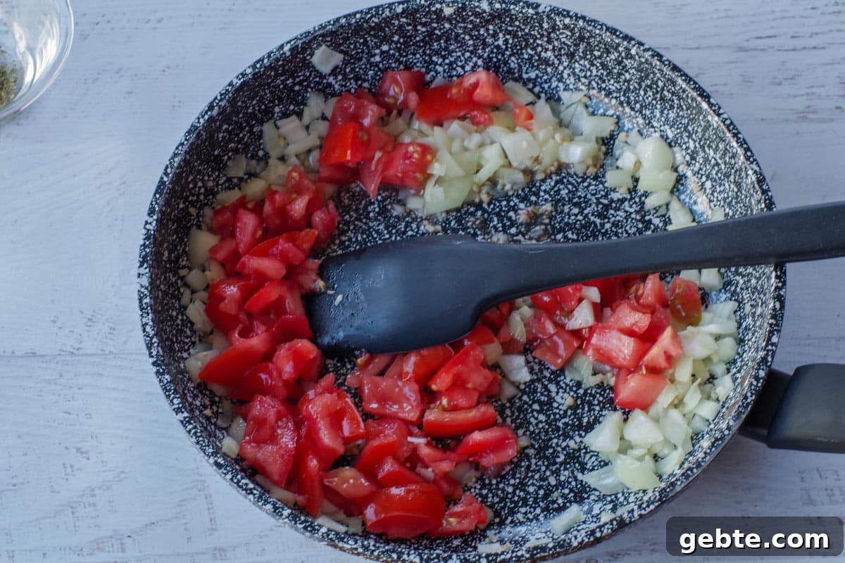 tomatoes added to frying pan with onion and garlic, with a black spatula