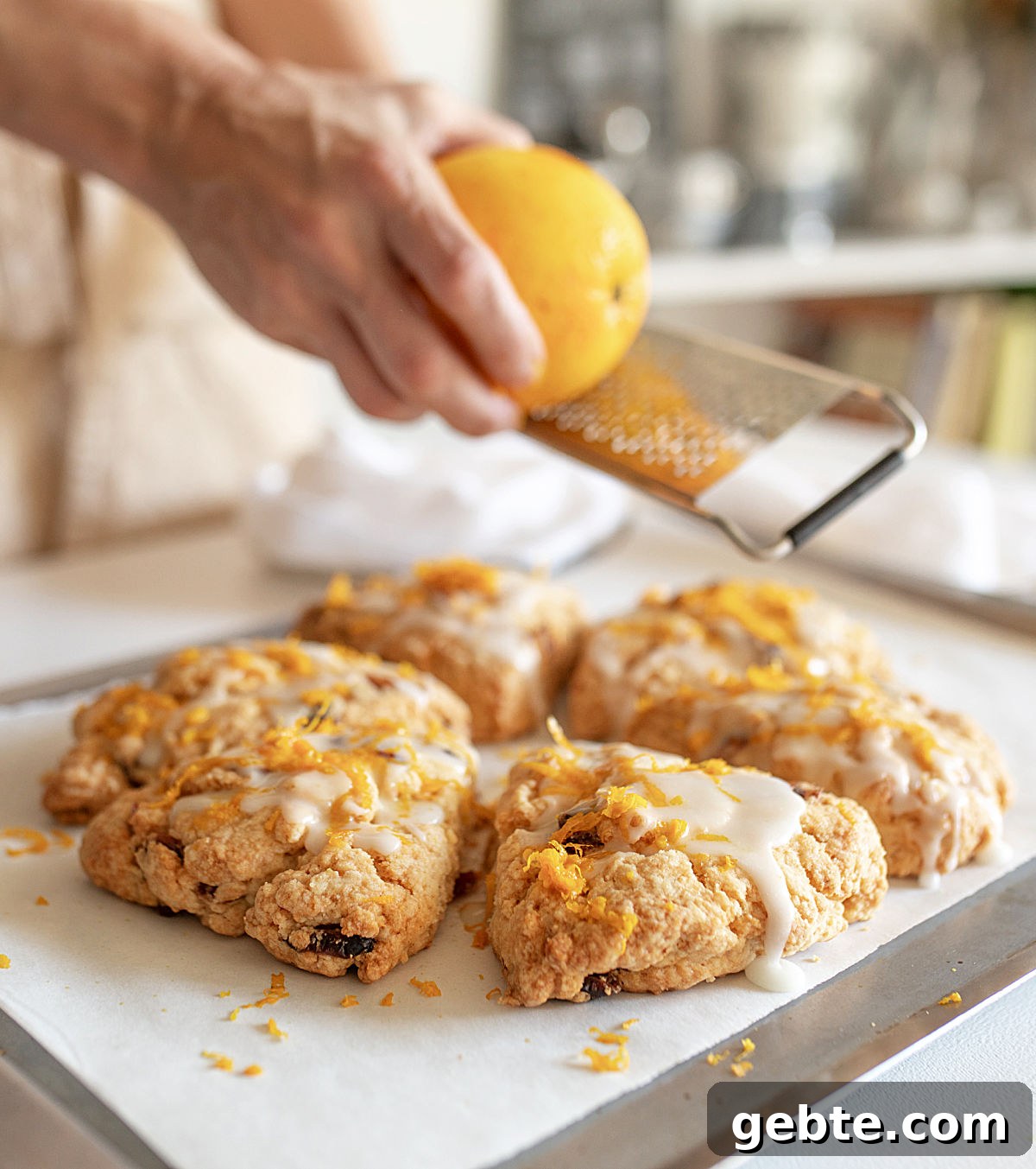 Grating orange peel over glazed scones on white paper and metal sheet pan.