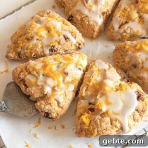Several triangle glazed scones with orange zest on white paper, metal cake server underneath