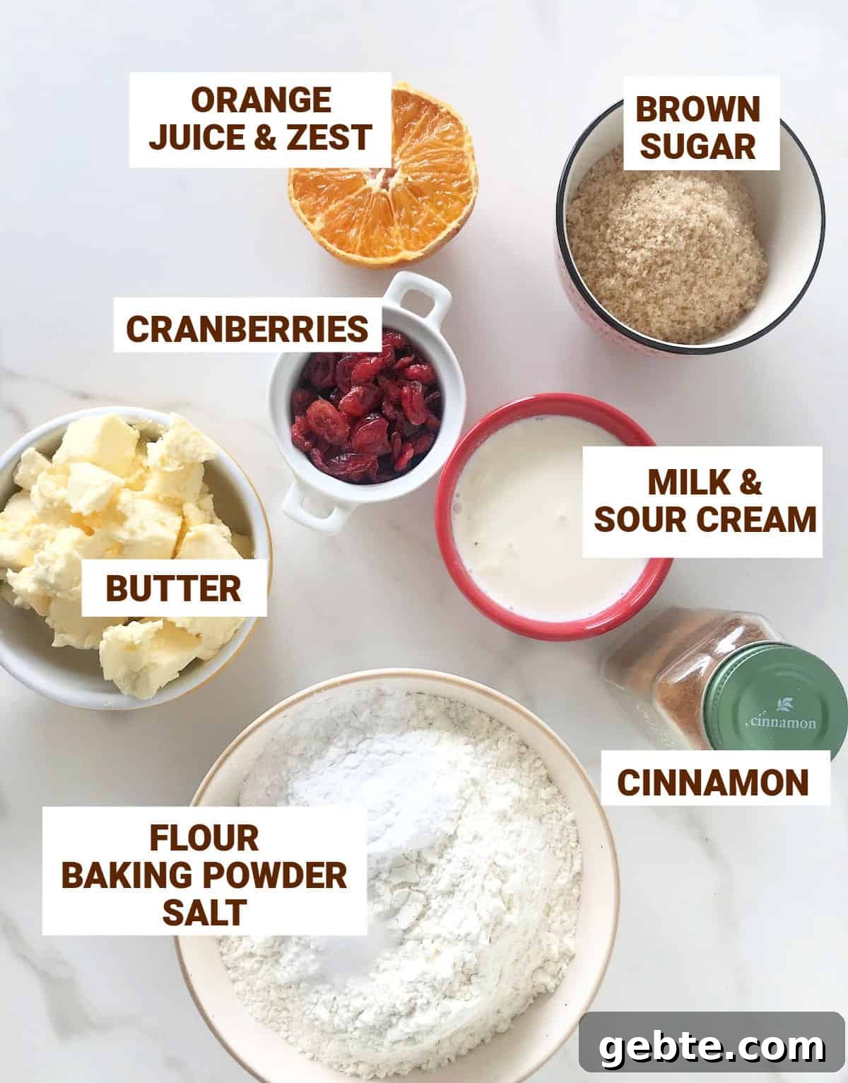 Ingredients for cranberry orange scones in bowls on a white surface including butter, cinnamon, flour mixture, brown sugar, milk and sour cream. .