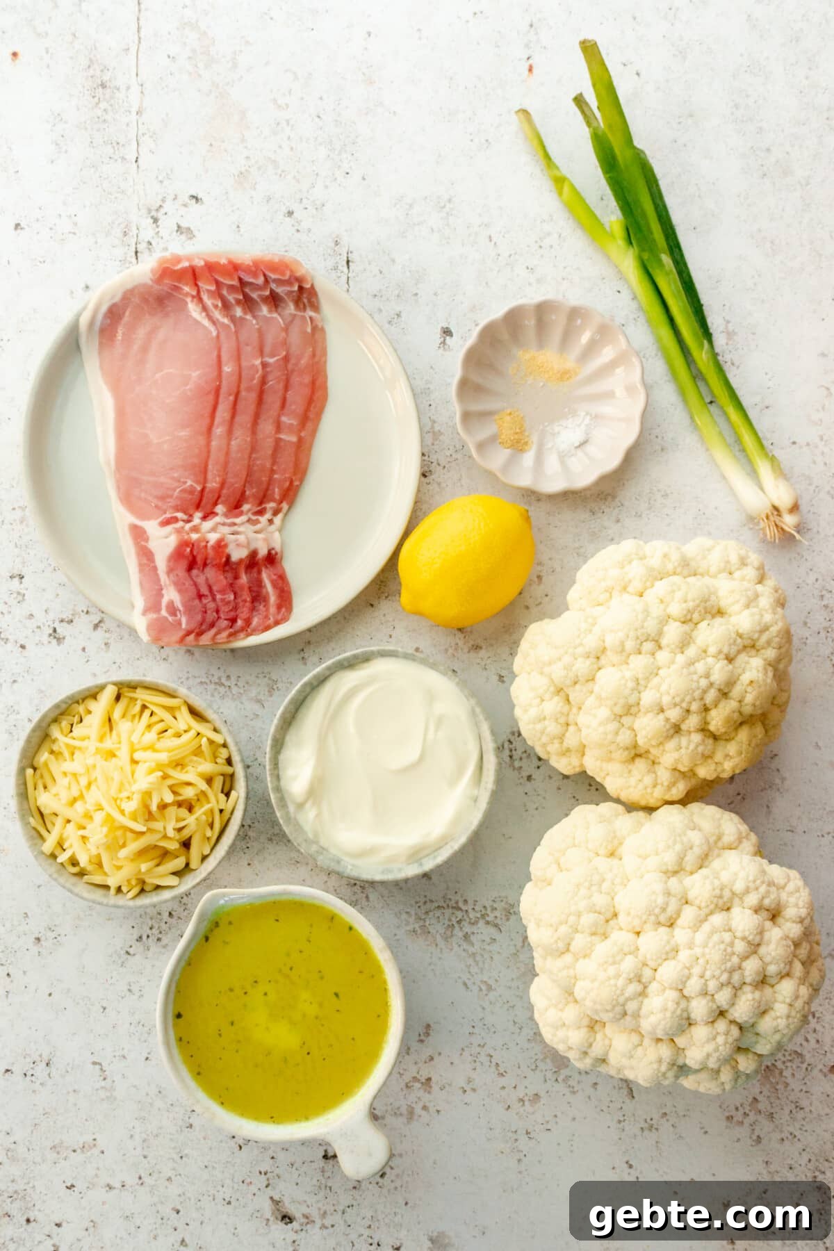 Ingredients for loaded cauliflower soup sit in a variety of bowls and plates on a light grey surface.