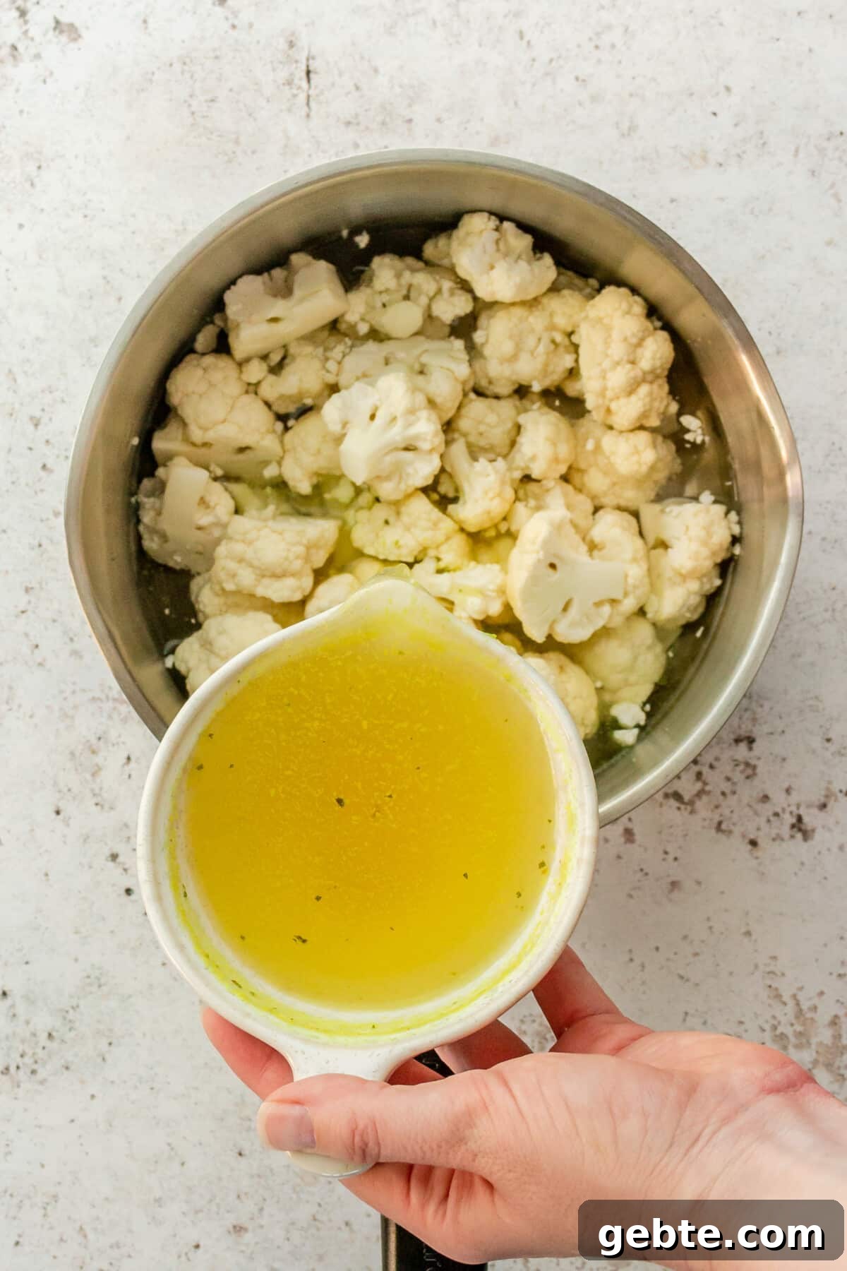 Broth is poured over cauliflower florets in a stainless steel saucepan on a light grey surface.