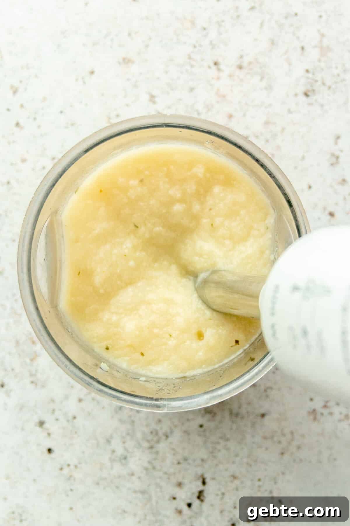 Cauliflower is blended in a immersion blender beaker on a light grey surface.