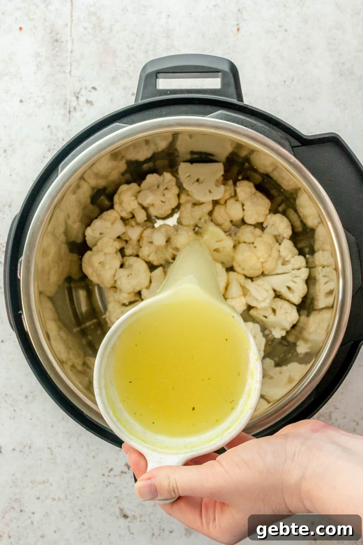 Broth is poured over cauliflower florets in an instant pot on a light grey surface.