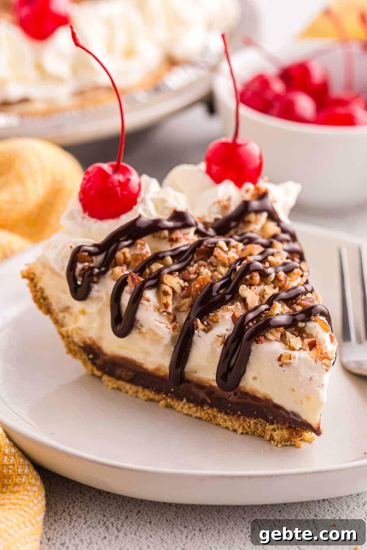 Slice of no bake banana split pie topped with whipped cream, chopped pecans, chocolate syrup, and cherries. The slice shows a graham cracker crust with creamy filling and a chocolate layer.
