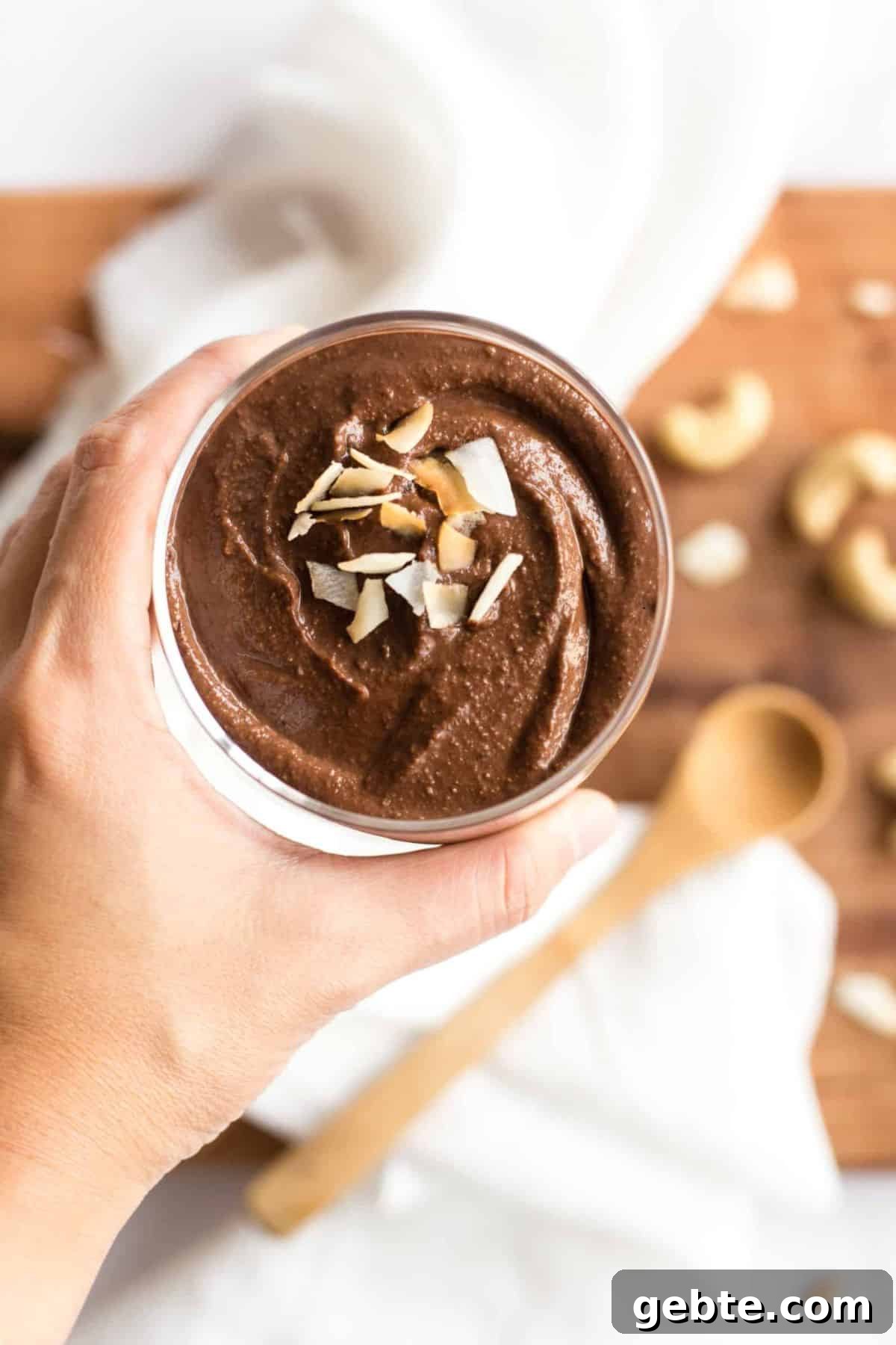 Vegan Cashew Chocolate Pudding Hand holding a cup of cashew chocolate pudding topped with coconut flakes.