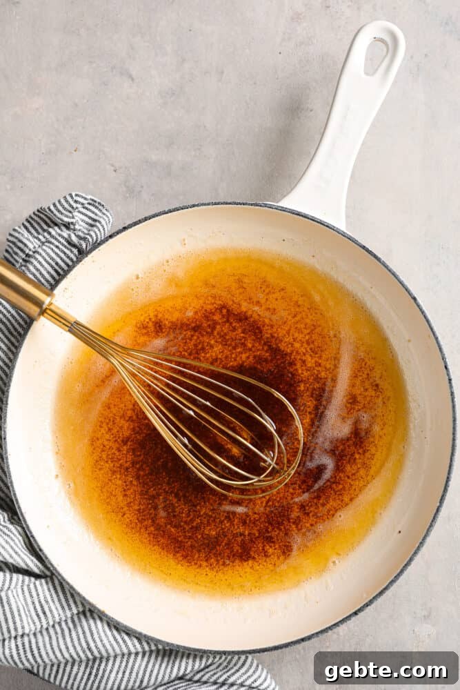 Learning how to brown butter will be your secret weapon in the kitchen! Easily done over the stove, butter slowly melts and releases a nutty toasted flavor that will enhance any sweet or savory recipe. Top view of browned butter in a white skillet. A gold whisk is inside the skillet with a blue and white striped towel next to the skillet.