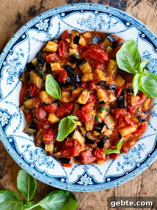 Sicilian Eggplant Caponata Recipe: Authentic Sweet-Sour Relish 9 Fried eggplant in tomato sauce with fresh basil in a blue and white bowl on a wooden surface.