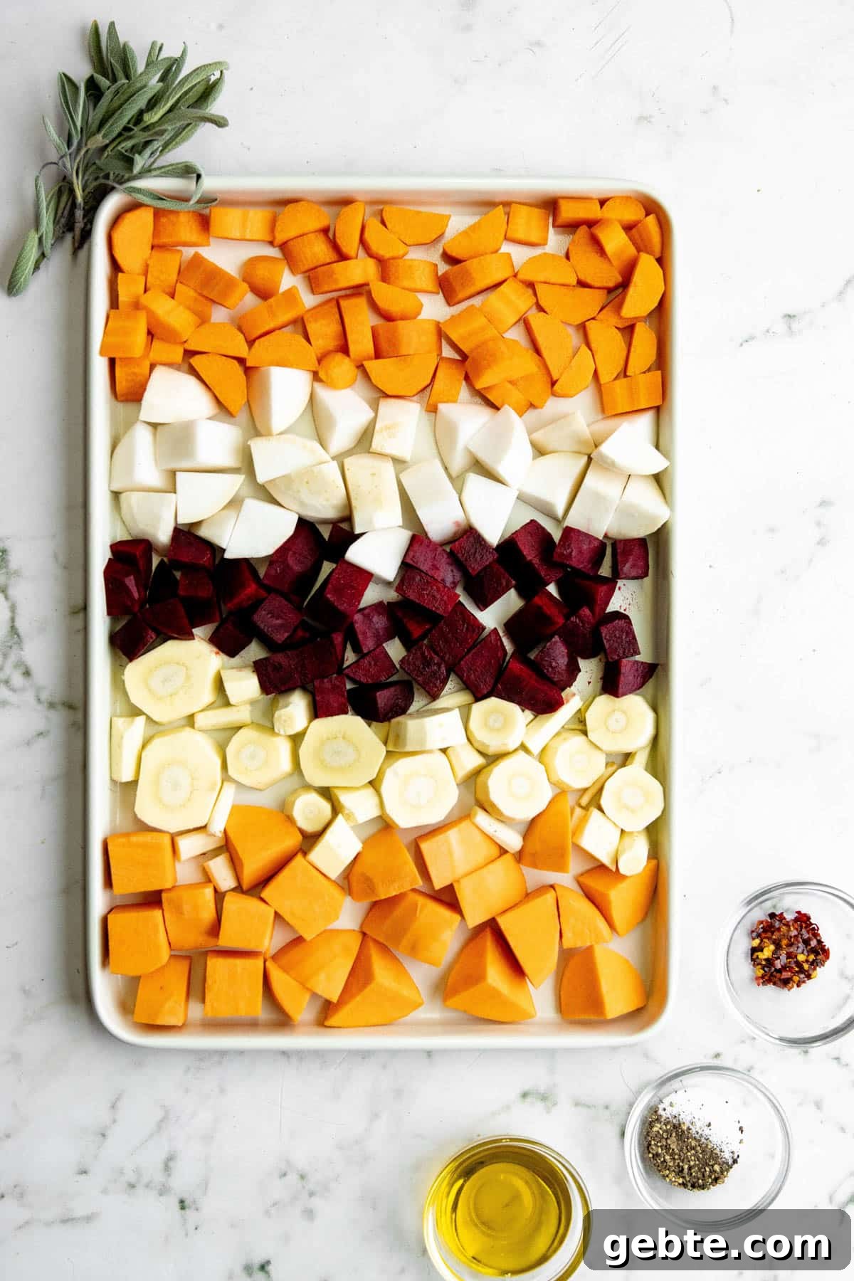 overhead photo of cubed root vegetables on a baking sheet, next to a bunch of sage and bowls of olive oil and seasonings