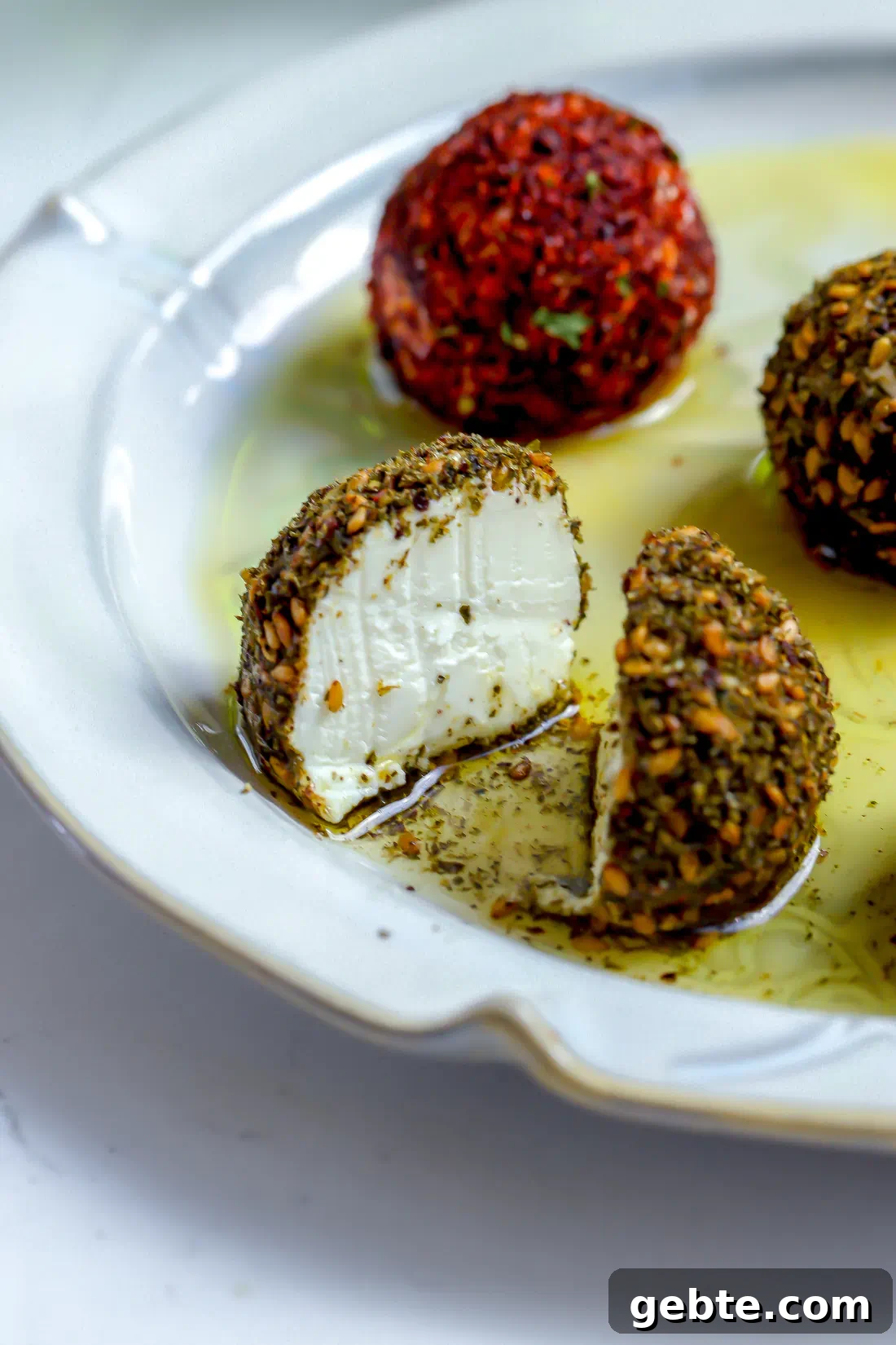 A close-up shot of a labneh ball coated in za'atar sliced in half to show the stark white, dense, and creamy yogurt cheese interior on a white plate with olive oil.