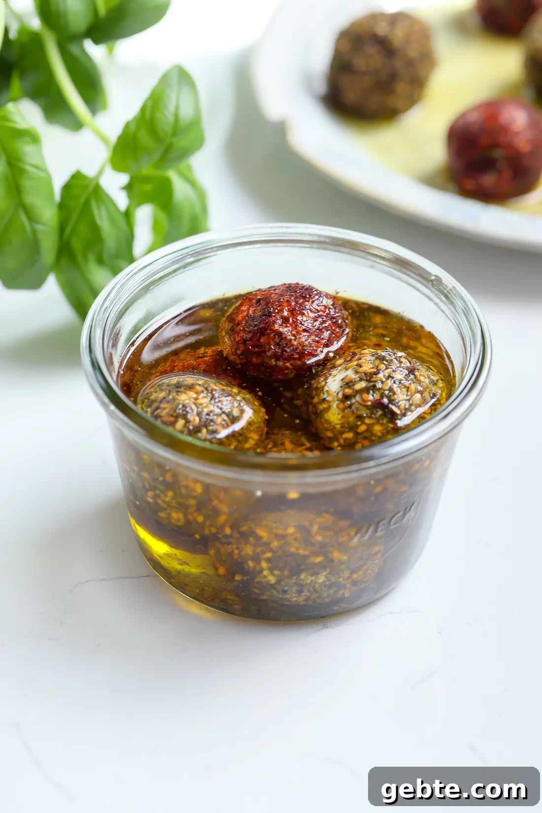 Top-down view of four labneh balls coated in red sumac and seedy za'atar, submerged in a glass jar of golden olive oil.