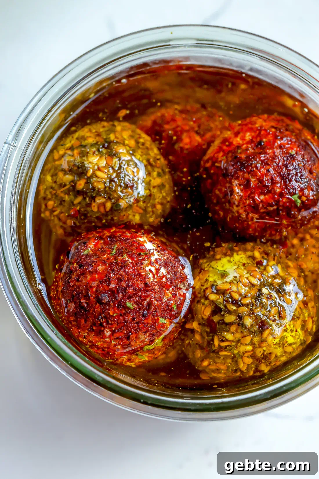 A glass jar of spiced labneh balls in olive oil on a white counter with fresh herbs and a serving plate in the background.