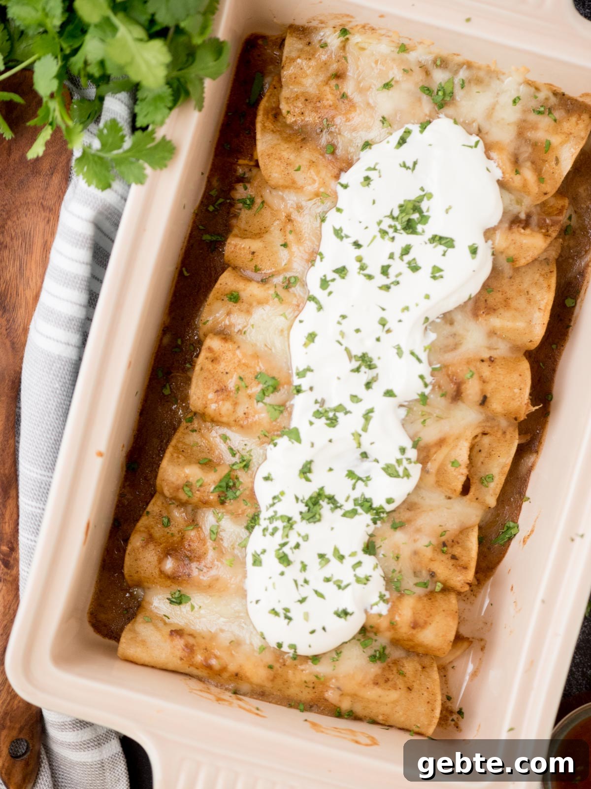 Authentic Red Chicken Enchiladas Recipe with Homemade Sauce 13 chicken enchiladas topped with sour cream and cilantro in a baking dish surrounded by serving utensils and fresh cilantro