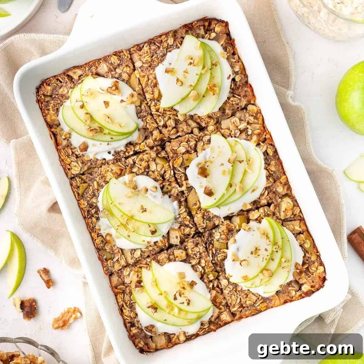 White rectangular dish of baked apple oatmeal, cut into six pieces and topped with yoghurt and apple slices.