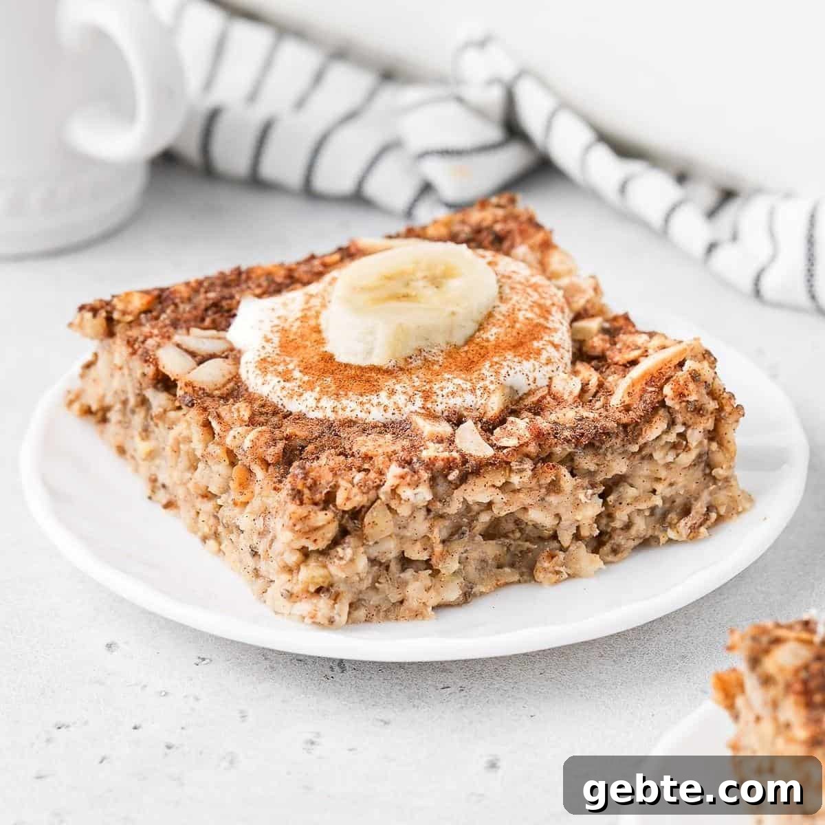 Piece of baked banana oatmeal on a round white plate, topped with some yoghurt, a slice of banana and cinnamon.