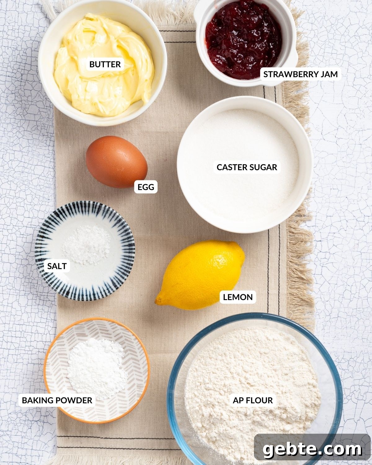 An overhead image of strawberry jam, butter, a fresh lemon, an egg, baking powder, caster sugar, and salt.
