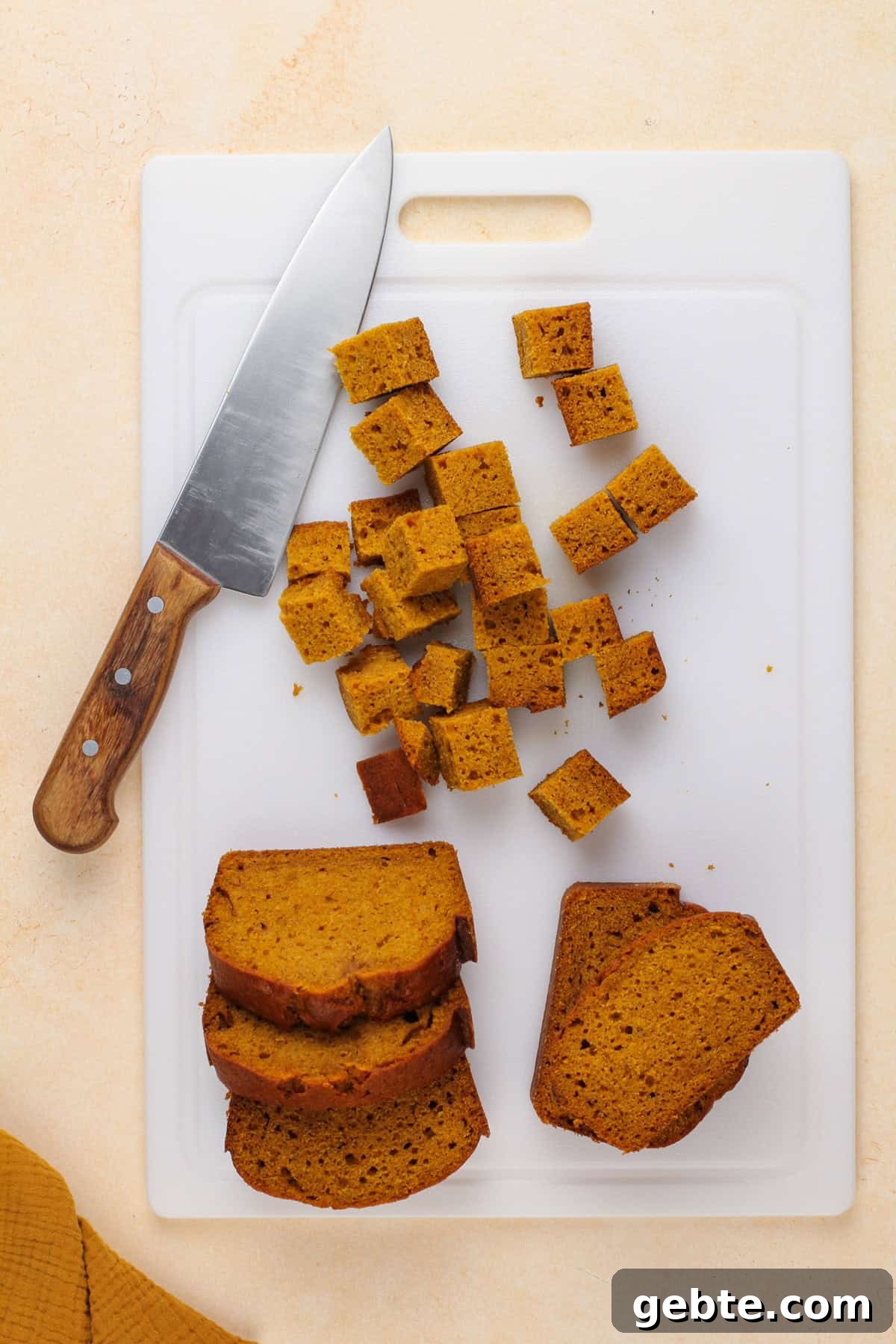 Pumpkin bread being cut into cubes on a white cutting board.