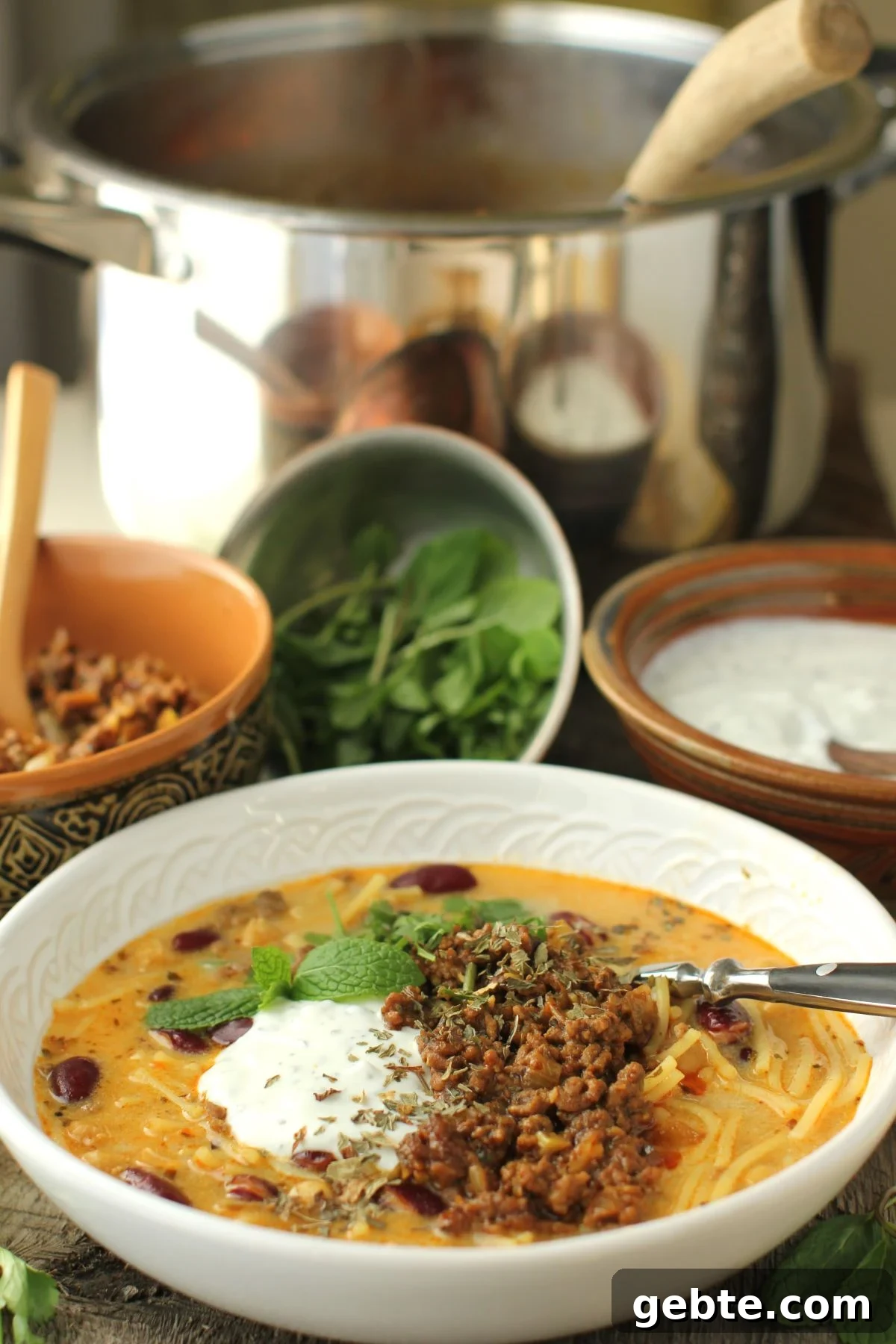 white bowl of aush soup with a blob of yogurt and spoonful of spiced meat on top. Soup pot behind and bowls of toppings.