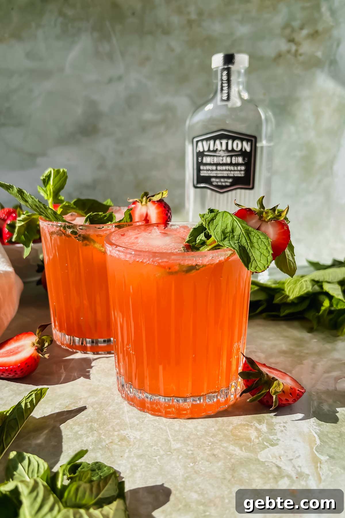 Strawberry Gin Smash: Refreshing Cocktail Recipe for Summer 2 two strawberry gin smash cocktails garnished with mint and strawberries with bottle of gin in background.