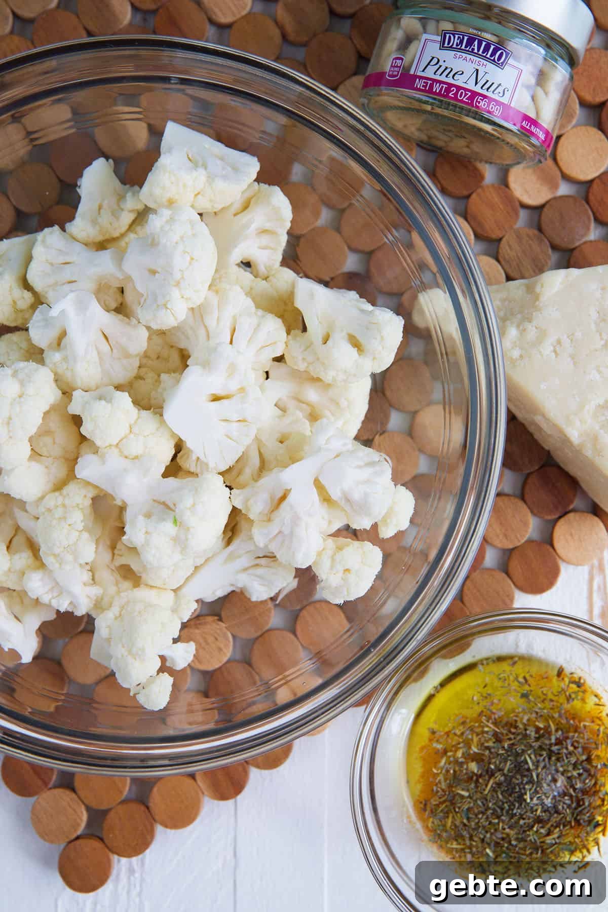 Crispy Oven-Roasted Cauliflower Recipe 3 A glass bowl filled with raw cauliflower is placed next to seasoned olive oil and pine nuts.