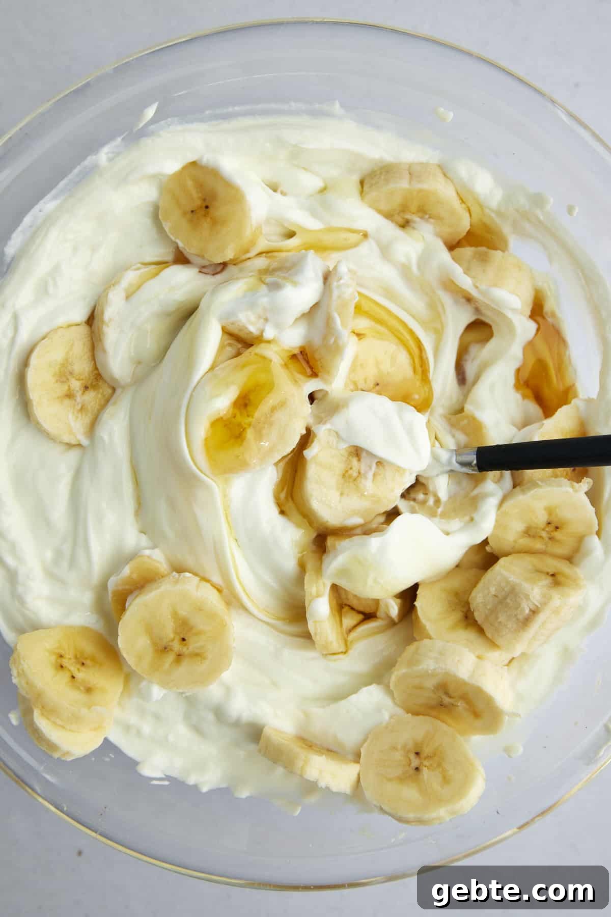 A mixture of Greek yogurt, honey, and banana coins in a mixing bowl with a spoon sticking out. 