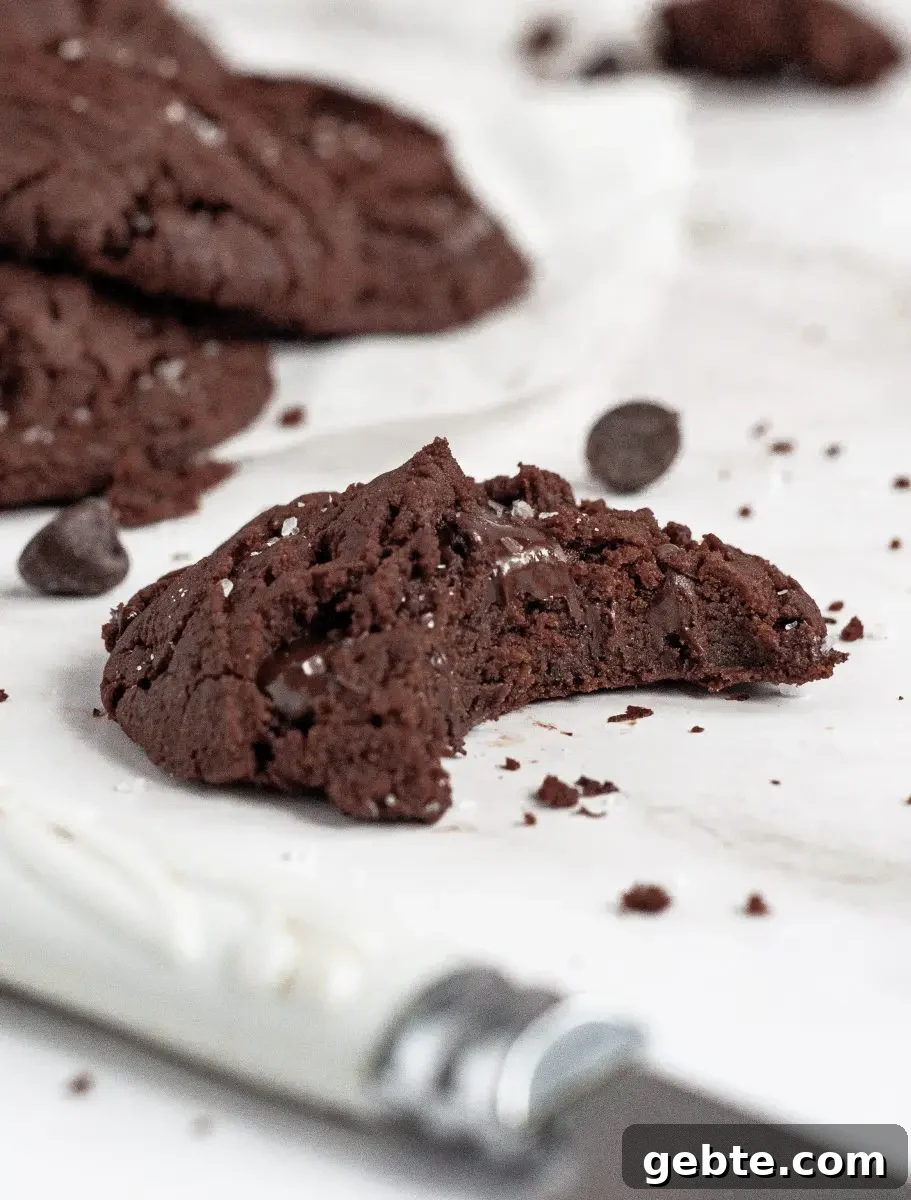 Gluten-Free Vegan Chocolate Mint Cookies Recipe 3 A dark brown chocolate mint cookies on a white piece of parchment paper. It has a large bite taken out of it and you can see the melted chocolate chips inside.