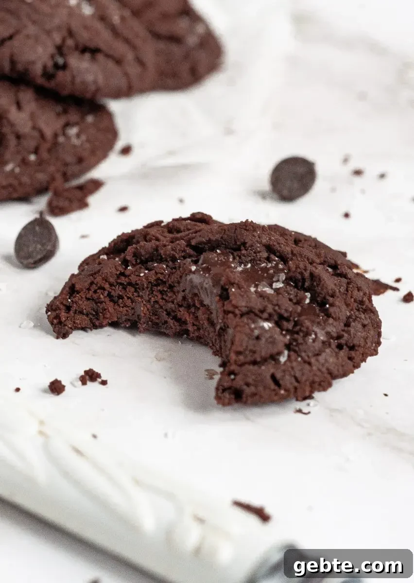 Gluten-Free Vegan Chocolate Mint Cookies Recipe 5 A dark brown chocolate mint cookies on a white piece of parchment paper. It has a large bite taken out of it and you can see the melted chocolate chips inside.