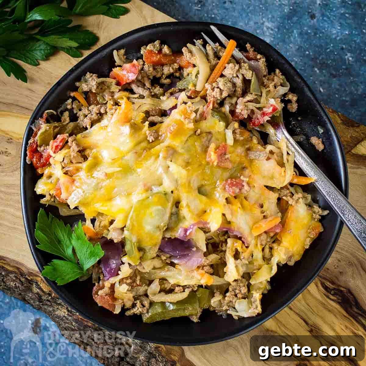 One-Skillet Stuffed Cabbage Casserole Recipe for Quick Weeknights 6 Close up overhead shot of cabbage casserole on a black plate with a silver fork on the side all on a wooden cutting board on a blue surface with herbs on the side.