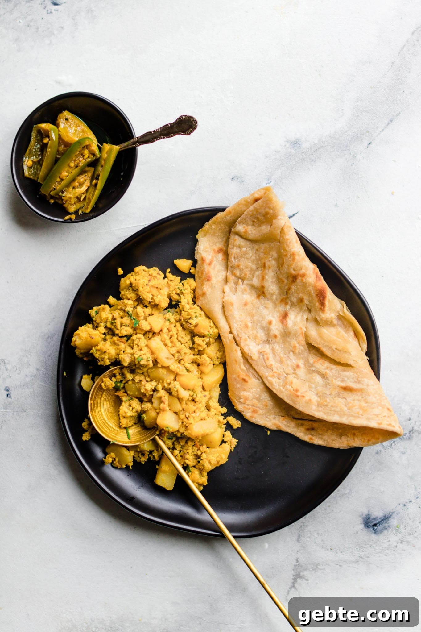 Scrambled Egg and Potato Curry with Paratha on a black plate with a side of achaar (mixed pickle)