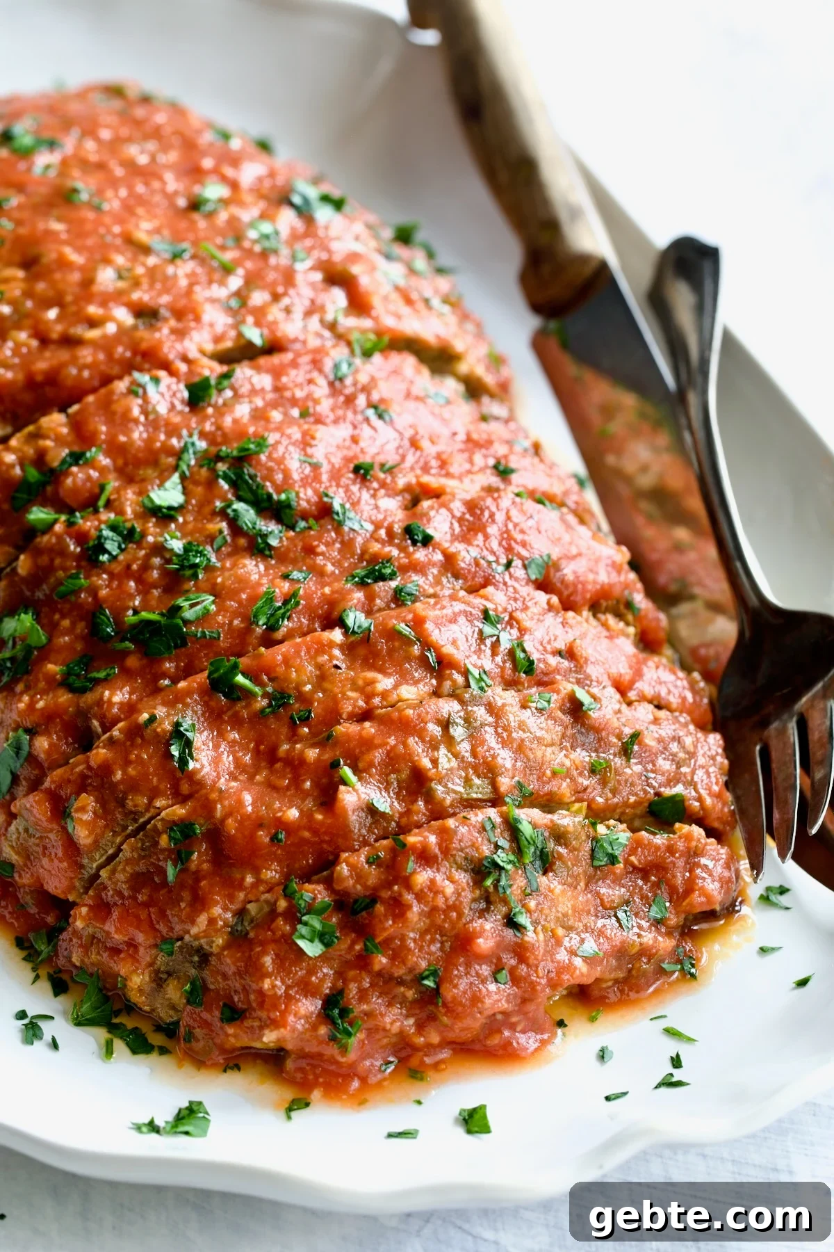 Italian Meatloaf on white platter garnished with fresh parsley with serving utensils.