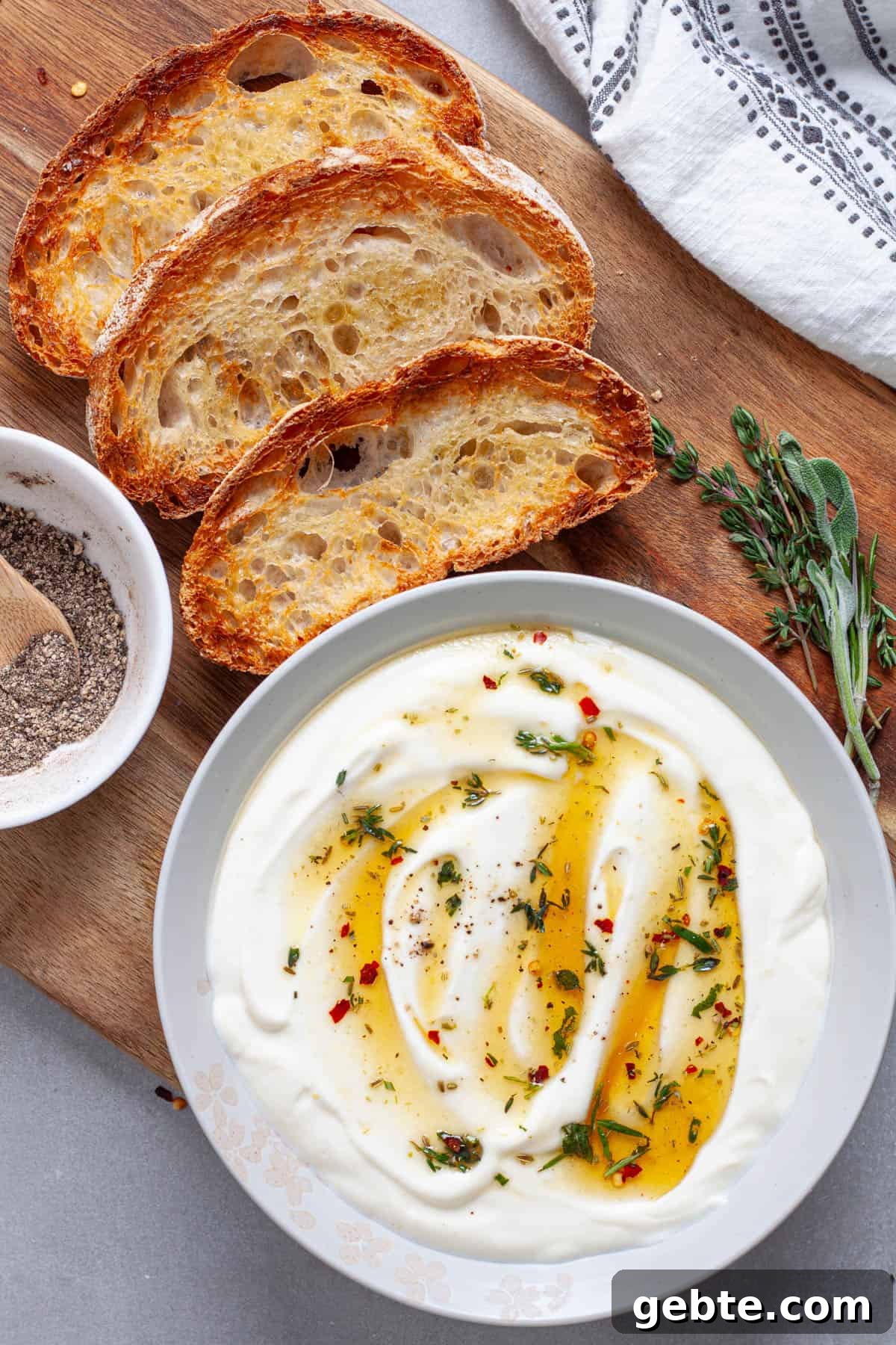 A bowl of whipped ricotta topped with honey and herbs on a cutting board with toasted slices of bread.