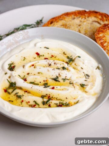 A serving bowl of whipped ricotta dip topped with honey and herbs on a plate with toasted bread.