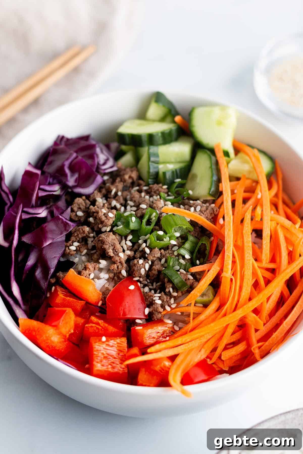 A white bowl with julienne carrots, red bell peppers, cabbage cucumbers and cooked ground beef. 