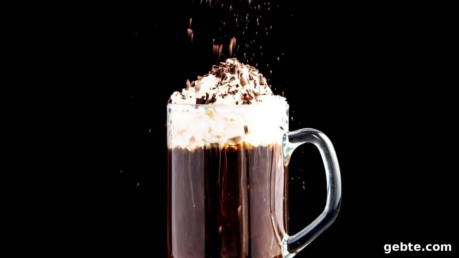 A glass mug of hot chocolate topped with whipped cream and chocolate shavings, with chocolate sprinkles falling from above against a black background.