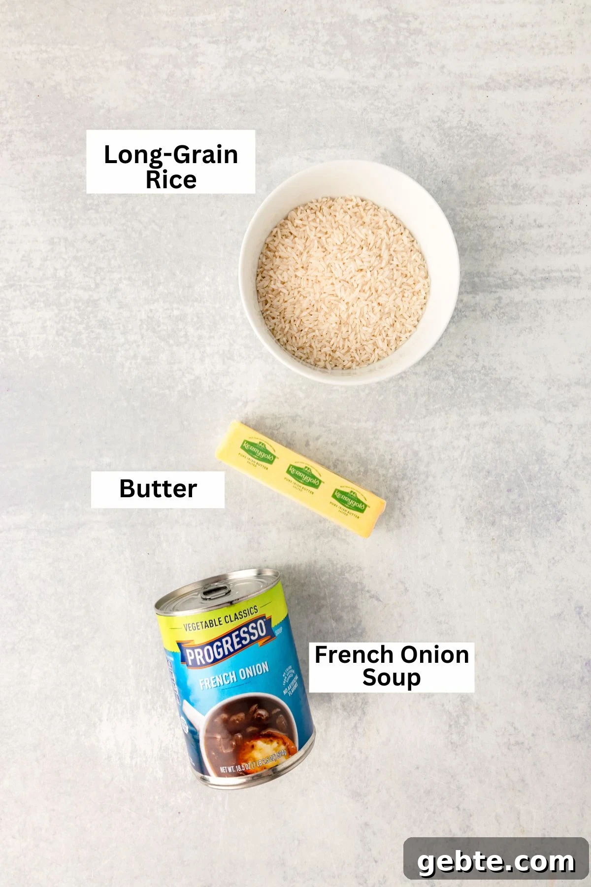 A bowl of long-grain rice, a stick of butter, and a can of French onion soup arranged on a light countertop, each labeled with its name.