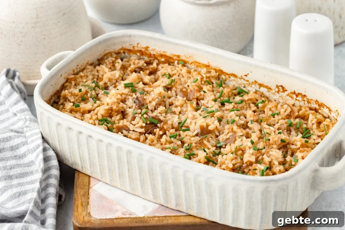 A white ceramic baking dish filled with baked rice, garnished with chopped chives, sits on a wooden board next to a striped kitchen towel and white salt and pepper shakers in the background.