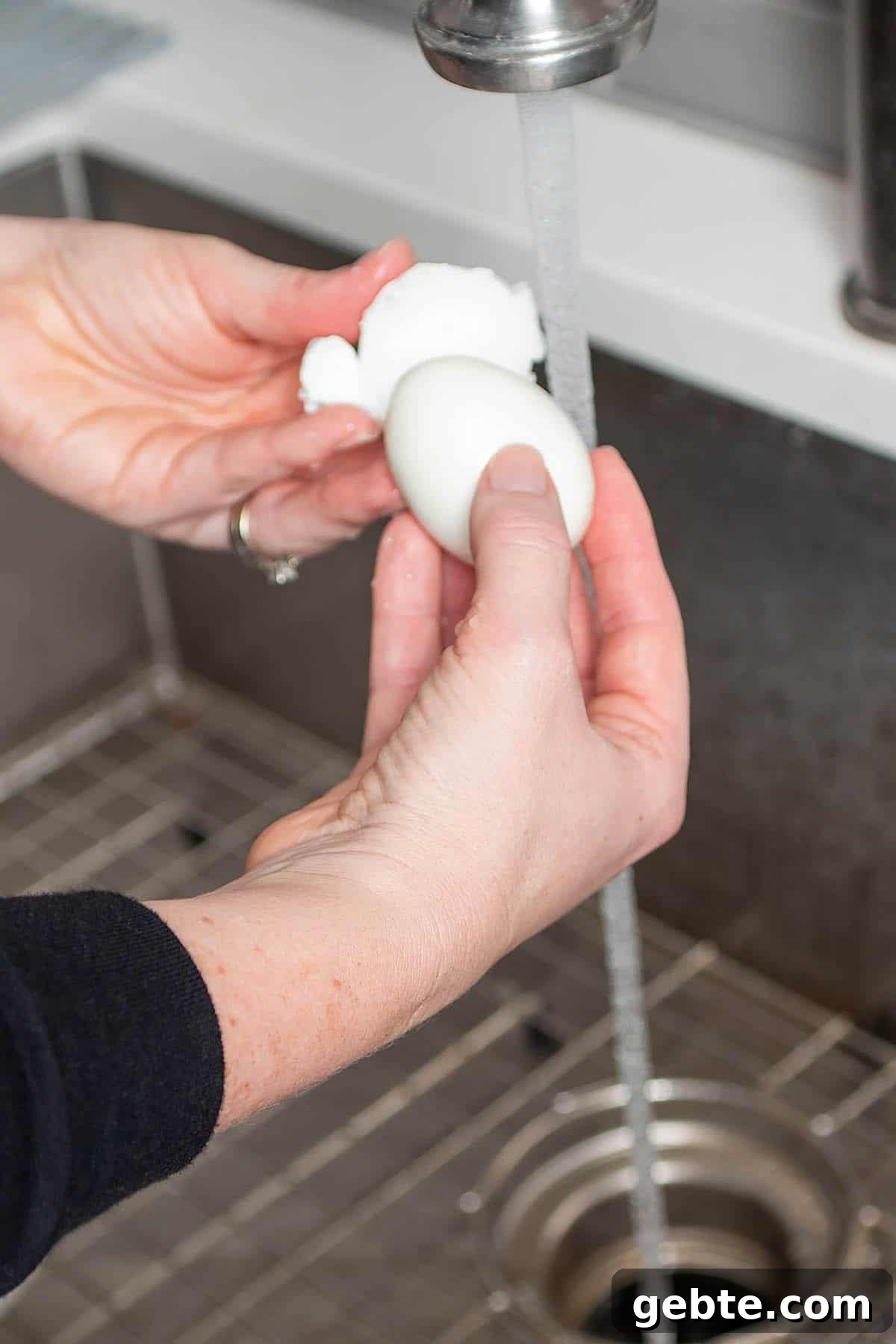 Two hands peeling an egg under running water.