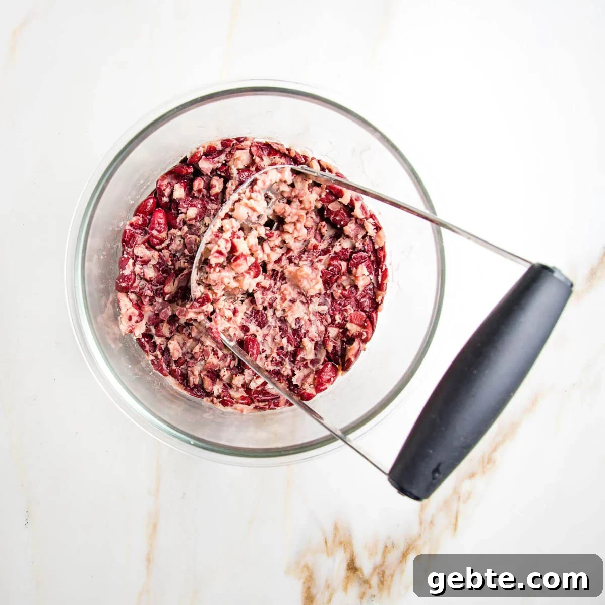 Potato masher squishing red kidney beans in a glass bowl.