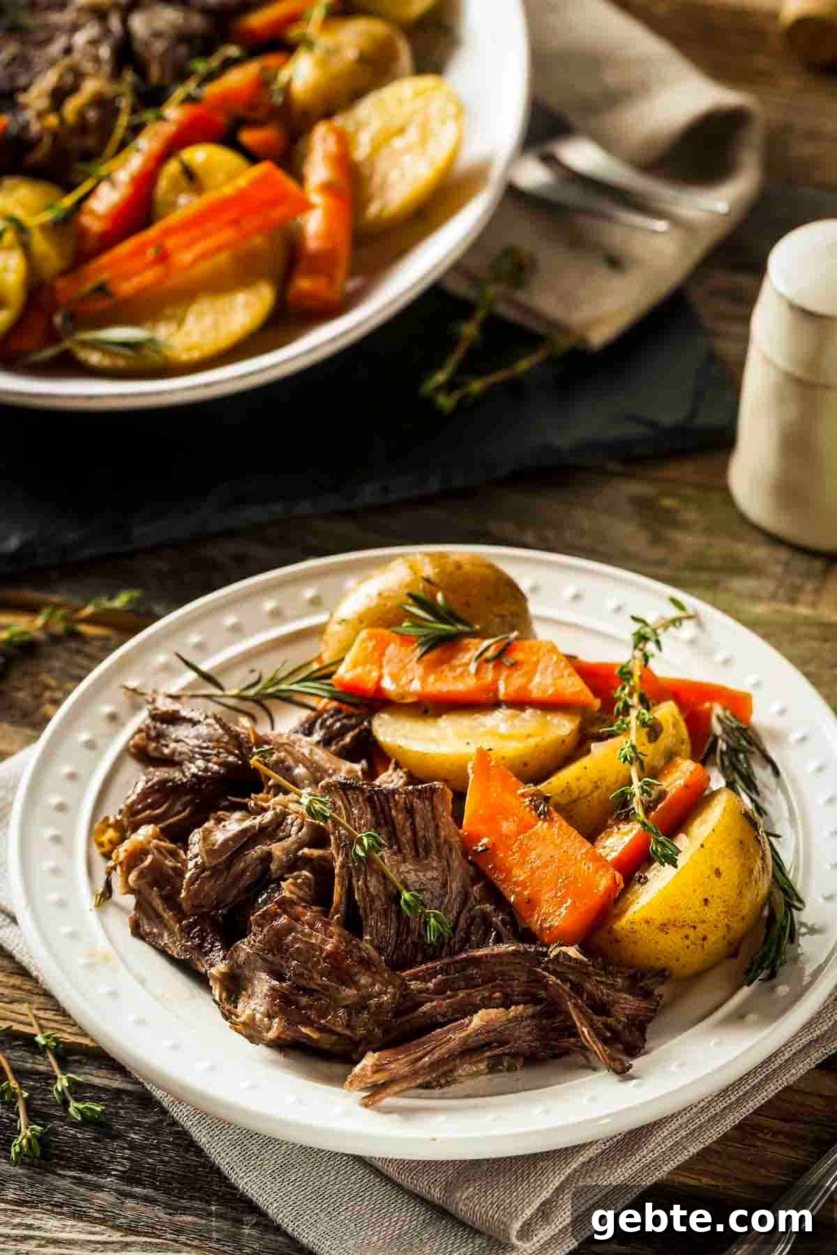Side shot of braised pot roast with carrots and potatoes on a white plate garnished with fresh rosemary.