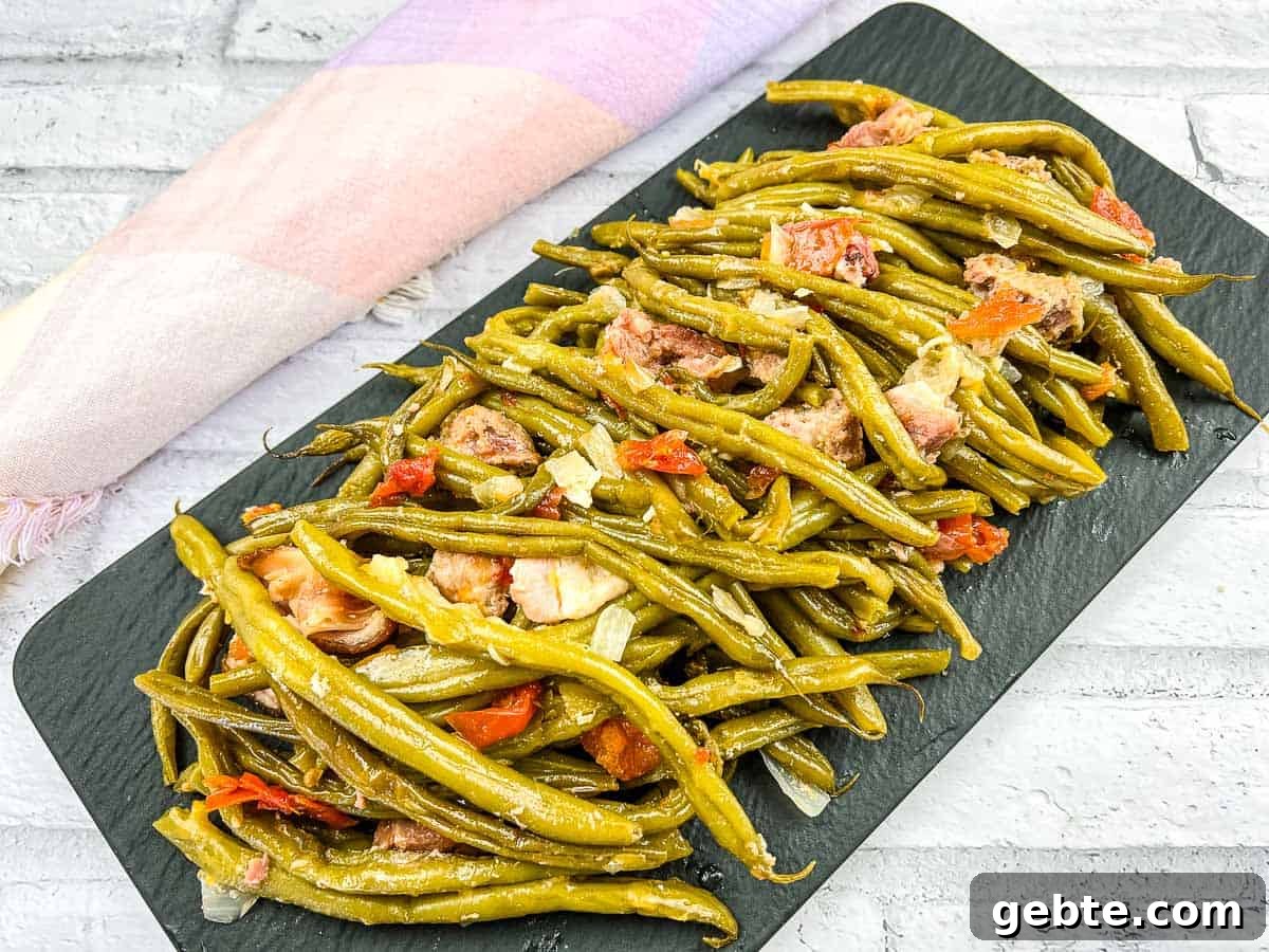 31 Slow Cooker Recipes to Walk In and Find Dinner Waiting 18 A plate of cooked Southern Green Beans with sliced sausages and tomato pieces on a white dish, placed near a sunflower.